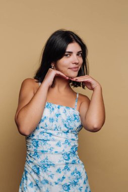 Siyah saçlı, gülümseyen ve elleri çenesinin altında şakacı pozlar veren bir kadın. Mavi çiçekli bir elbise giyiyor ve sıcak bej bir arka planda duruyor..