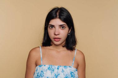 Düz bir bej arka planın önünde duran bir kadın tarafsız bir ifade sergiliyor. Açık renk çiçekli bir elbise giyiyor ve omuz hizasında siyah saçları var..