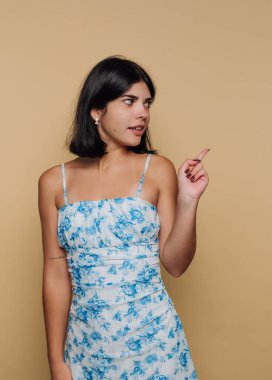 Genç bir kadın açık mavi çiçekli bir elbise içinde, tek eliyle sıcacık bir bej arka plan üzerinde şakacı bir şekilde duruyor..