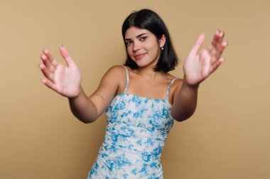 Kısa siyah saçlı genç bir kadın mavi çiçekli bir elbise giyer ve kollarını açar, sıcak bir şekilde gülümser. Tarafsız kahverengi arka plan onun neşeli yüz ifadesini güçlendirir..