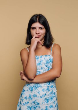 Genç bir kadın, çiçekli bir elbise giyerek kollarını kavuşturarak güvenle ayakta duruyor. Düşünceli ve rahat görünüyor, sıcak bir bej arka planda duruyor..