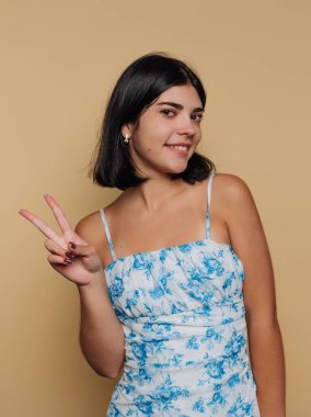 Genç bir kadın kameraya poz verirken kendinden emin bir şekilde gülümsüyor. Mavi çiçekli bir elbise giyiyor ve eliyle barış işareti gösteriyor. Tarafsız arka plan onun neşeli ifadesini vurguluyor..