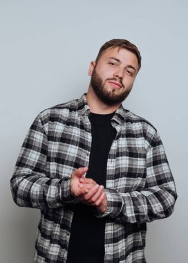 Sakallı genç bir adam siyah gömlek ve ekoseli düğme giyiyor. Elleri birarada, tarafsız bir arkaplana karşı kendinden emin bir tavır sergiliyor..