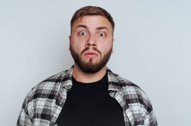 Bir adam evde dururken şaşırmış bir yüz ifadesi sergiler. O, siyah bir tişörtün üzerine ekose bir gömlek giyer. Sade arka plan tepkisini vurguluyor ve odaklanmasını sağlıyor..