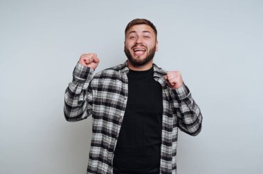 Siyah tişörtü ve kareli gömleği olan bir adam arka planda duruyor. Heyecanla gülümsüyor, kutlama için yumruklarını kaldırıyor, mutluluk duygusunu iletiyor..