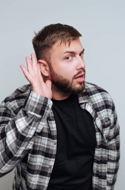 Sakallı genç bir adam, söylenenlere ilgi göstererek elini kulağına dayıyor. O, siyah bir tişörtün üzerine flanel bir gömlek giyiyor, basit bir gri arkaplan üzerine kurulmuş..