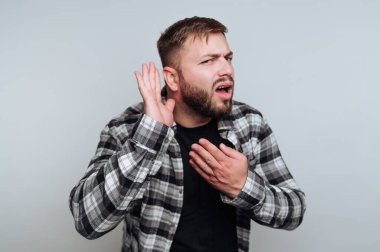 Ekose gömlekli genç bir adam şaşırmış görünüyor, bir kulağını avuçlarken bir elini göğsüne koyuyor. O, dikkatlice dinlemeye çalışıyor gibi görünüyor. Işık arka planı ifadesini güçlendiriyor..