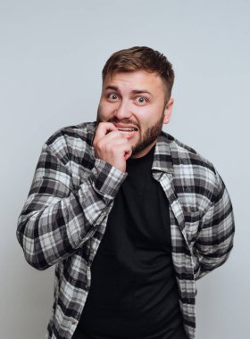 Sakallı ve ekose gömlekli bir adam endişeli görünüyor, arka planda dikilirken tırnaklarını yiyor. İfadesi, sıradan bir ortamda huzursuzluk ve gerilim yayıyor..