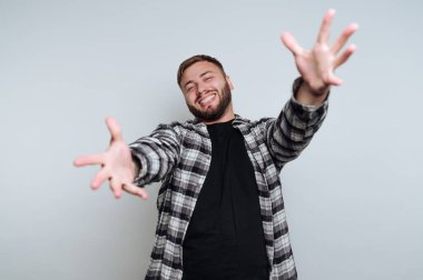 Neşeli genç bir adam kollarını genişçe açarken geniş bir şekilde gülümser, neşe ve açıklık hissi taşır. Sıradan bir ortamda siyah bir tişört ve kareli bir gömlek giyiyor..