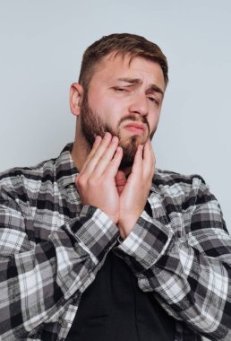 Bir adam içeride duruyor, belli ki rahatsız bir şekilde çenesini tutuyor. Ekose bir gömlek giymiş ve bir ağrı ya da endişe duyuyor gibi görünüyor..