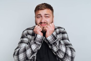 Neşeli bir suratı olan genç bir adam yumruklarını çenesinin yakınında sıkıyor, heyecan ya da gerginlik duyguları sergiliyor. Tarafsız bir arkaplanı olan basit bir kapalı alanda duruyor..