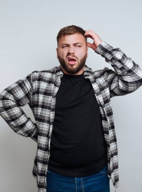 Kareli gömlek ve siyah tişört giyen bir adam kafası karışmış, açık gri bir arkaplanda dikilirken kafasını kaşıyor, şaşkınlığını ifade ediyor..