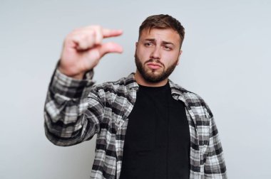 Ekose gömlek giyen genç bir adam parmaklarını birbirine yakın tutuyor sanki küçük bir nesnenin boyutunu gösteriyor gibi. Ciddi bir ifadesi var ve düz bir arkaplana karşı duruyor..