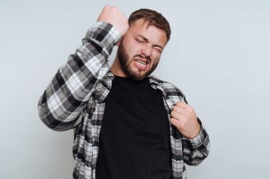 Genç bir adam kutlama için yumruğunu kaldırıyor, siyah bir tişörtün üzerine ekose bir gömlek giyiyor. Mekan, basit, hafif bir arka planda, bir zafer anı ya da mutluluk anıdır..