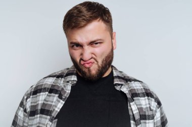 Siyah bir tişörtün üzerine ekose gömlek giyen bir adam komik bir ifade sergiliyor. Basit bir beyaz arkaplana karşı poz verir, neşeli bir anı yakalar ve eğlencesini iletir..