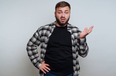 Bir adam şaşkın bir ifade ile ayağa kalktı ve bir kareli gömlek ve siyah tişört giymiş el kaldırdı. Açık gri bir arka plana karşı kapalı alanda, gündelik güveni somutlaştırıyor..