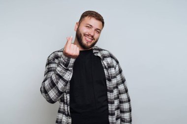 Sakallı genç bir adam gülümsüyor ve parmaklarıyla kalp hareketi yapıyor. O, ekose bir gömleğin altına siyah bir gömlek giyiyor, basit bir gri arka fonun önünde duruyor..