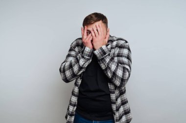 Genç bir adam düz bir arka planda duruyor, elleriyle yüzünü örtüyor. Vücut dili şaşkınlık ya da korku gösteriyor. Kareli gömlek ve koyu renk kot pantolon giyiyor..