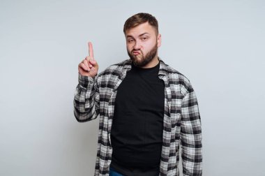 Genç bir adam siyah bir gömlek ve ekose bir fanila giyer, bir parmağını şakacı bir ifadeyle kaldırır. Basit bir arkaplanın önünde rahatça poz veriyor..