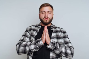 Sakallı genç bir adam elleriyle sakince meditasyon yapıyor. Ekose bir gömlek giyiyor ve sakin bir yerde, gözleri kapalı, iç huzura odaklanmış..