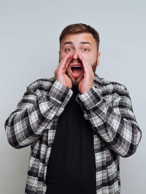 Kareli gömlek giyen bir adam heyecanını yüksek sesle ifade eder, ellerini ağzına atar. Sade bir arka plana karşı duruyor, neşeli tepkisini gösteriyor..