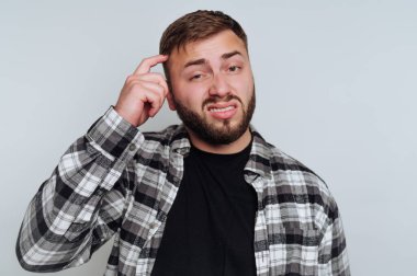 Genç bir adam sade bir arka planın önünde şaşkın şaşkın bakıyor. Eli başının üzerinde duruyor ve siyah bir tişörtün üzerine ekose bir gömlek giyiyor..