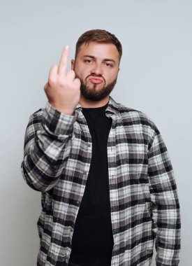 Genç bir adam düz bir arkaplanda duruyor, siyah bir gömlek ve kareli bir fanila giyiyor. Yüzünde ciddi bir ifadeyle orta parmağını kaldırıyor..