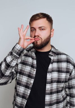 Siyah bir tişörtün üzerine ekose gömlek giymiş genç bir adam içeride duruyor. Kendine güveni tam bir jest yapıyor. Tarz ve kendine güvenini minimalist bir ortamda sergiliyor..