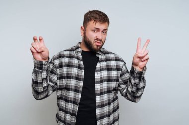 Siyah bir tişörtün üzerine ekose gömlek giymiş genç bir adam düz bir arkaplanda duruyor. İki eliyle barış işareti yapıyor ve neşeli bir ifadesi var..