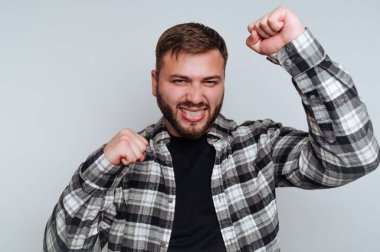 Ekose gömlek giyen genç bir adam kolları havada ve yumruklarını sıkarak mutluluğunu ifade ediyor. Neşeli yüz ifadesi basit bir iç mekanda zafer duygusu ya da kutlama hissi veriyor..