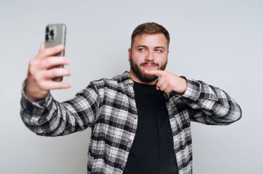 Genç bir adam selfie çekerken gülümseyen hafif bir arkaplan karşısında duruyor. Kareli bir gömlek giyiyor ve neşeli görünüyor, çenesini gösteriyor, anın tadını çıkarıyor..