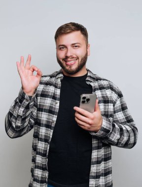 Sakallı ve arkadaş canlısı ifadeli genç bir adam stüdyoda duruyor. Bir elinde akıllı bir telefon, diğer elinde ise kendine güven ve pozitiflik gösteren bir işaret tutuyor..