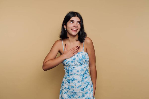 Молодая женщина стоит перед обычным фоном в синем цветочном платье. Она тепло улыбается, прикладывая руку к сердцу. Атмосфера приятная и уютная.