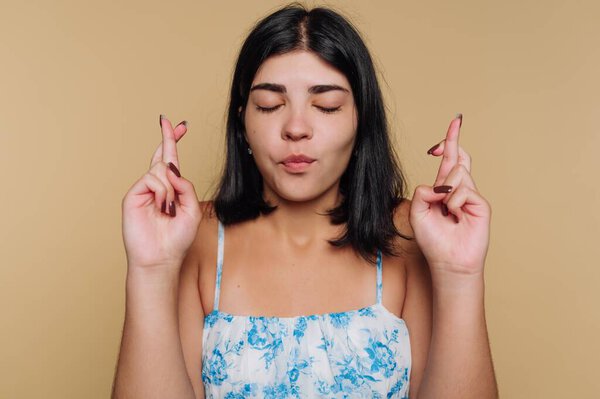 Молодая женщина скрещивает пальцы и закрывает глаза, выражая надежду и ожидание. Она носит цветочный топ и стоит на теплом фоне, создавая спокойную атмосферу..