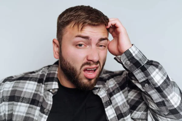 Kısa saçlı genç bir adam, elini başının üstüne koyarak karışık bir yüz ifadesi sergiliyor. Siyah bir gömlek ve bir ekose ceket giyer, açık bir arka fonda durur..