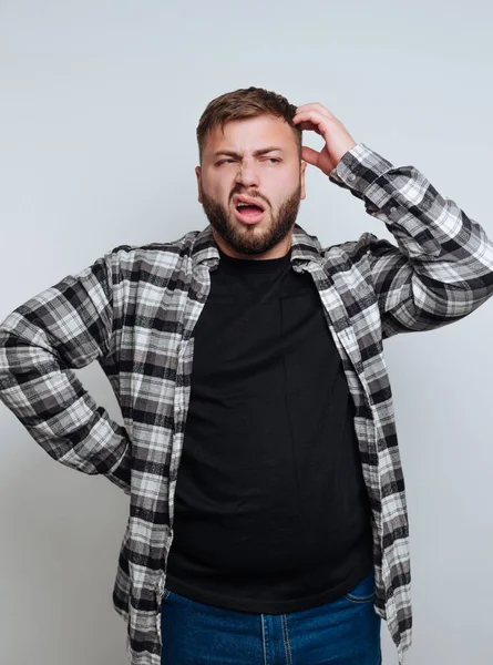 Kareli gömlek ve siyah tişört giyen bir adam kafası karışmış, açık gri bir arkaplanda dikilirken kafasını kaşıyor, şaşkınlığını ifade ediyor..