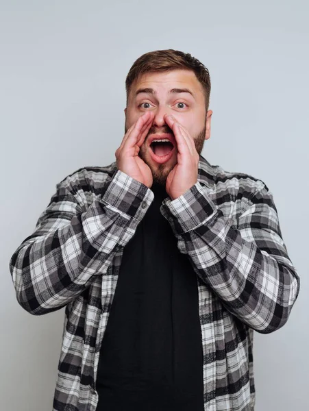 Kareli gömlek giyen bir adam heyecanını yüksek sesle ifade eder, ellerini ağzına atar. Sade bir arka plana karşı duruyor, neşeli tepkisini gösteriyor..