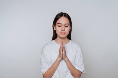 Bir kadın, elleri göğsünün önünde, gözleri kapalı, sakin ve minimalist bir alanda meditasyonu kucaklayarak sessizce ayakta durur..