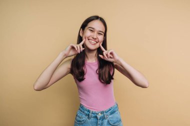 Genç bir kız bej bir duvarın önünde duruyor, yanaklarının yanında neşeli parmak hareketleri yaparken parlak bir şekilde gülümsüyor. Pembe bir bluz ve kot şort giyiyor, neşeli bir anı yakalıyor..