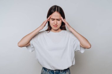 Genç bir kadın gözleri kapalı ve elleri başının üstünde duruyor. Stres ya da acı belirtileri gösteriyor. Beyaz bir gömlek ve kot şort giyiyor. İlişkili bir an yaşanıyor..