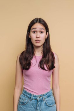 Genç bir kız şaşırmış bir ifadeyle ayakta duruyor. Pembe kolsuz bir bluz ve kot şort giyiyor. Arka plan sıcak bir bej, basit ama çarpıcı bir kontrast oluşturuyor..