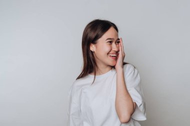 Uzun saçlı genç bir kadın, yüzünün yarısını şakacı bir şekilde eliyle örterken parlak bir şekilde gülümsüyor. Sade arka plan onun neşeli ifadesini ve rahatlamış ruh halini vurguluyor..