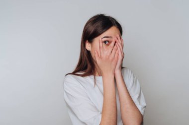 Genç bir kadın iyi ışıklandırılmış bir alanda duruyor, elleriyle yüzünü kapatıyor, kameraya bakarken şaşkınlık ve utangaçlık karışımı bir şey gösteriyor..