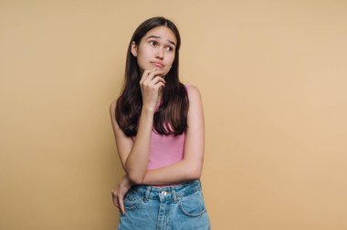 Genç bir kız rahat bir poz veriyor, çenesinde bir parmak ile düşünceli görünüyor. Pembe bir bluz ve kot şort giyiyor, yumuşak pastel arka planda..