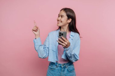 Genç bir kadın pembe bir arka planın önünde duruyor, bir elinde akıllı bir telefonla gülümsüyor ve işaret ediyor. Üzerinde mavi çizgili gömlek ve kot pantolon var..