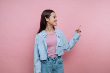 Açık mavi bir gömlek ve pembe bir üst giyen gülümseyen genç bir kadın, neşeli bir an gösteren yumuşak pembe bir arka plana karşı mutlu bir şekilde onun tarafını işaret ediyor..