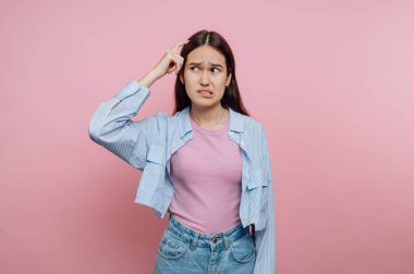 Genç bir kadın pembe bir duvarın önünde duruyor, kafasını kaşıdığında kafası karışmış görünüyor. Açık mavi bir kot ceket ve pembe bir bluz giyiyor. Sıradan bir görünüşü var..