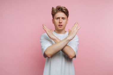 Genç bir adam pembe bir arka planın önünde duruyor, kollarıyla çapraz bir jest yapıyor. Şaşırmış ya da endişeli görünüyor, bir ifade ve duygu anı yakalıyor..