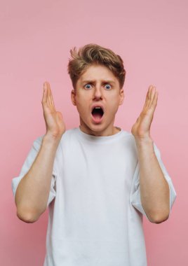 Açık kahverengi saçlı genç bir adam beyaz bir tişört giyer ve şok olmuş bir ifade gösterir. Pembe bir arka plana karşı şaşkınlıkla ellerini kaldırır çarpıcı bir görsel etki yaratır..