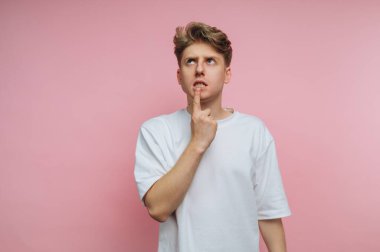 Genç bir adam pembe bir duvarın önünde duruyor, düşünceli bir ifade sergilerken parmağını dudaklarına değdiriyor. Basit beyaz bir tişört giyiyor ve düşüncelere dalmış gibi görünüyor..
